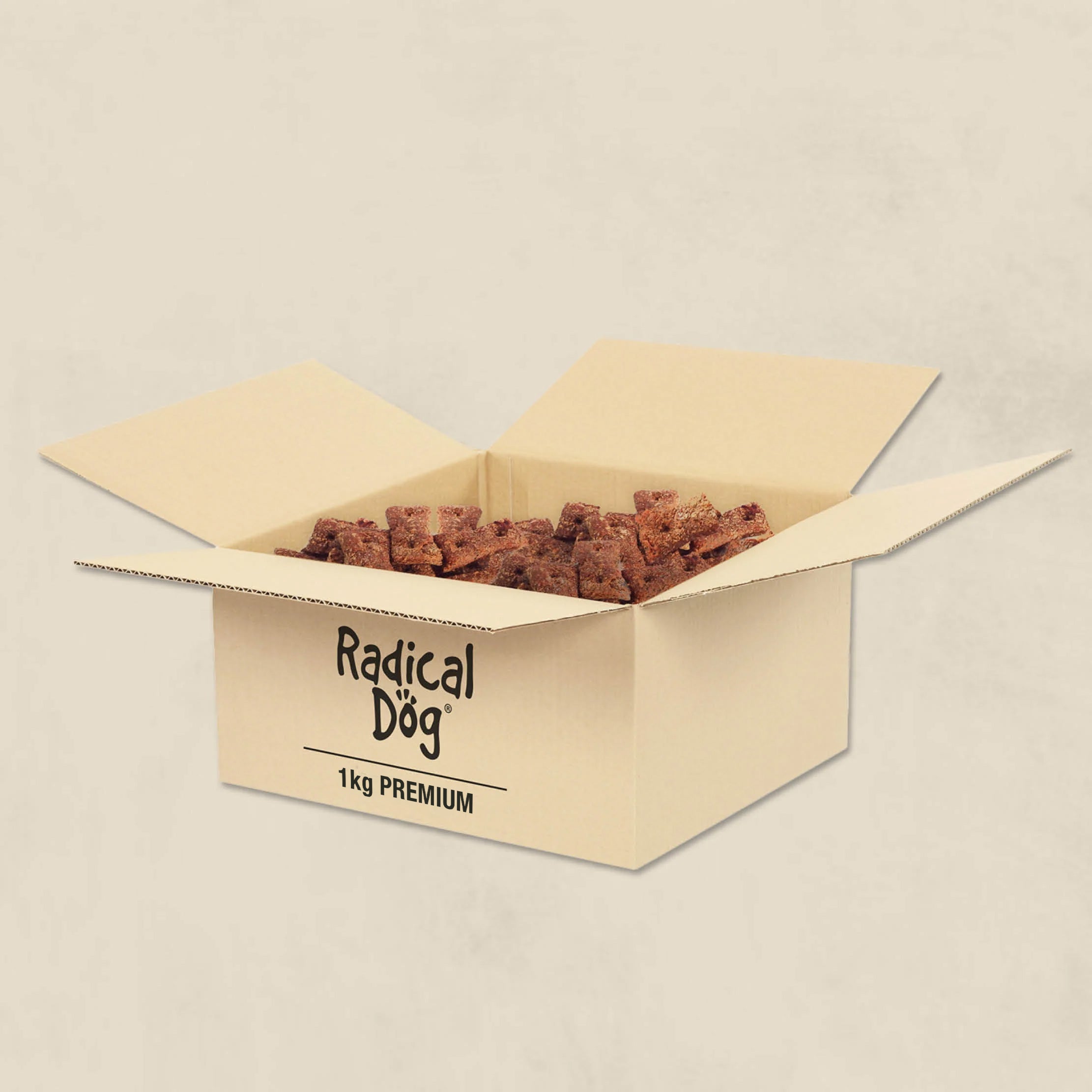 An open cardboard box filled with brown dog treats, with the brand name 'Radical Dog' and '1kg PREMIUM' printed on the front, set against a light beige textured background.